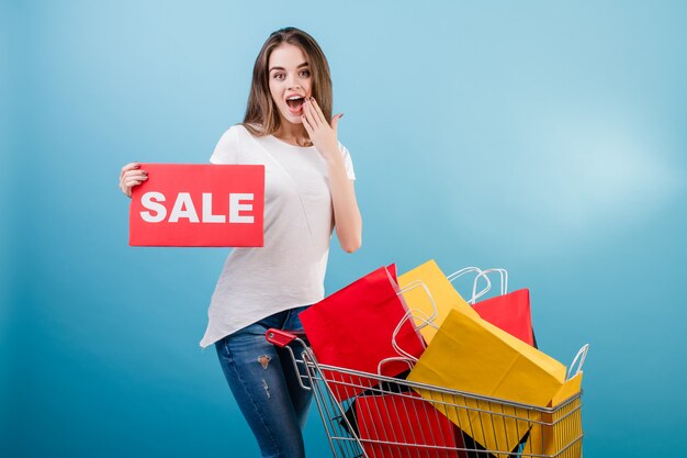 Brunette woman with shopping cart full of colorful red and yellow paper bags and sale sign isolated over blue