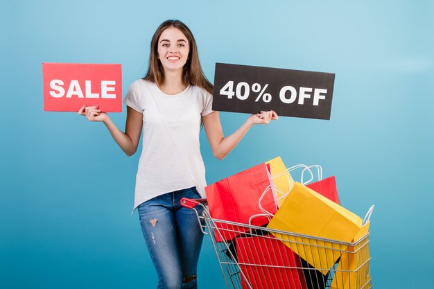 Brunette woman with shopping pushcart full of colorful red and yellow paper bags and 40% sale sign isolated over blue