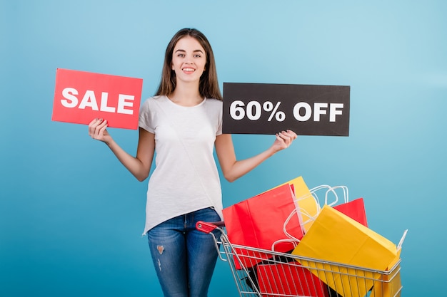 Brunette woman with shopping pushcart full of colorful red and yellow paper bags and 60% sale sign isolated over blue