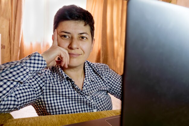 Photo brunette woman with short hair in check shirt sits in front of laptop screen, pensively looks aside and smiles slightly. work from home, freelancer.