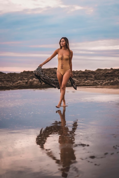 A brunette woman with a swimsuit and a pareo walking along the beach in summer in the cloudy sunset