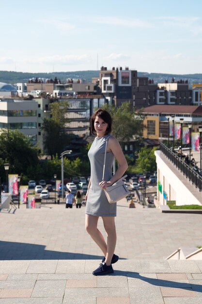 A brunette woman with a wide smile stands on a bridge against the background of the city sunny