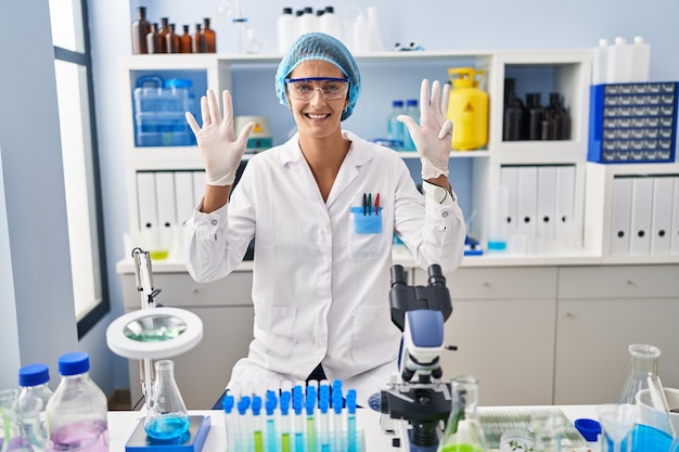 Brunette woman working at scientist laboratory showing and pointing up with fingers number nine while smiling confident and happy