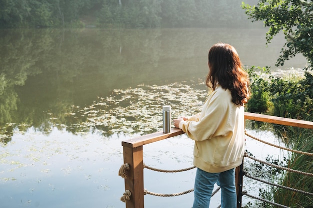 Donna castana in felpa con cappuccio gialla con tazza termica rilassante vicino al lago al mattino presto