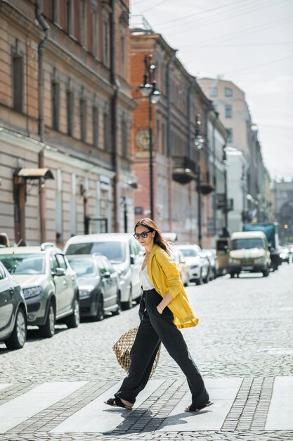 Brunette woman in a yellow jacket and black pants, sunglasses and bag string bag walk on the street.