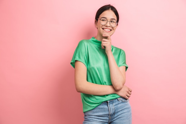 brunette young girl wearing round eyeglasses touching her chin and smiling at front isolated over pink wall