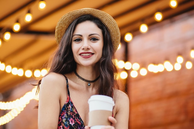 Brunette young lady in summer straw hat and dress, having make-up, holding paper cup with hot drink, having rest at outdoor cafeteria, posing at camera with smile, demonstrating her white teeth.