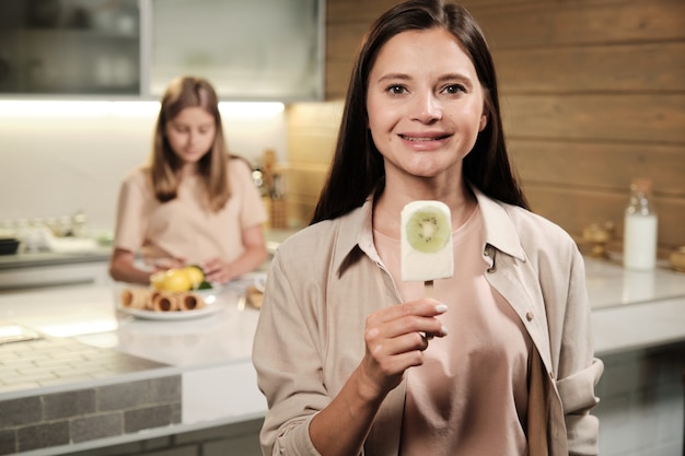 Brunette young smiling woman showing you homemade eskimo icecream with fresh kiwi while eating it in front of camera in the kitchen