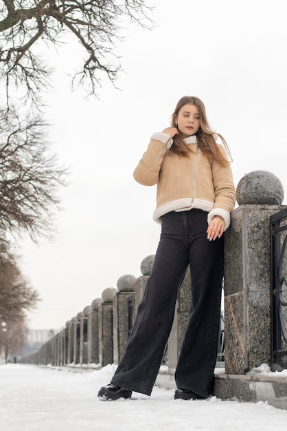 Brunette young woman in black jeans and beige sheepskin coat stands pensively in winter park Winter look for every day