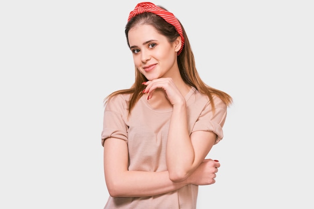Brunette young woman smiles positively wearing casual outfit and looking to the camera with hand on chin isolated on white wall People emotions