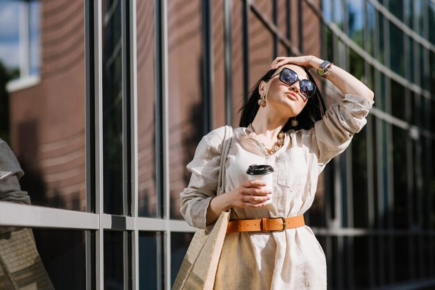 Фото Брюнетка молодая женщина в темных очках и сумке с кофе гуляет по городу. образ жизни портрет женщины