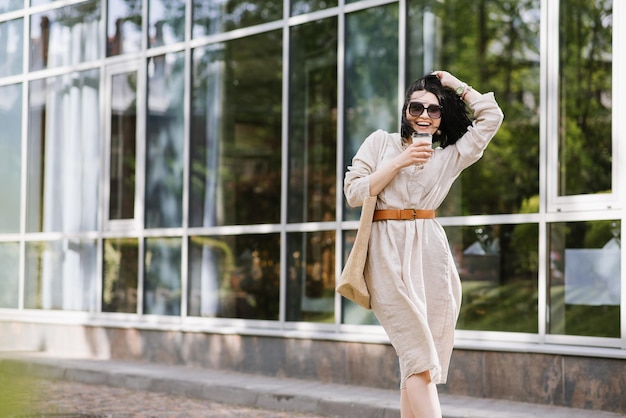 Giovane donna bruna con occhiali da sole e borsa che tiene caffè a piedi in città. ritratto di stile di vita di donna