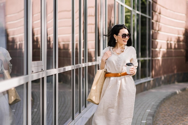 街を歩いてコーヒーを保持しているサングラスとバッグを持つブルネットの若い女性。女性のライフスタイルの肖像画