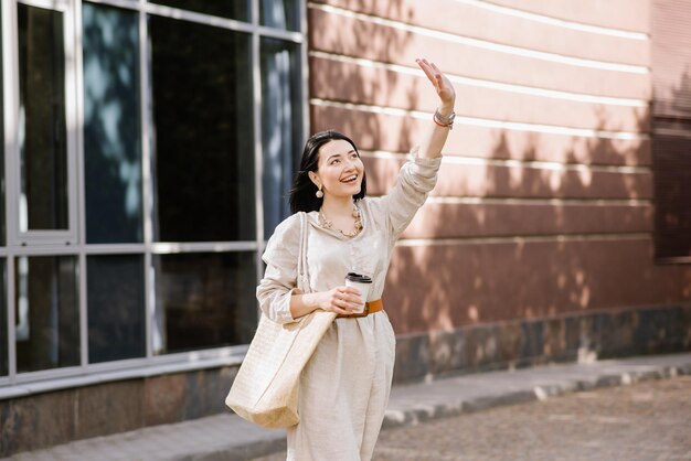 Photo brunette young woman with sunglasses and bag holding coffee walking in the city. lifestyle portrait of woman