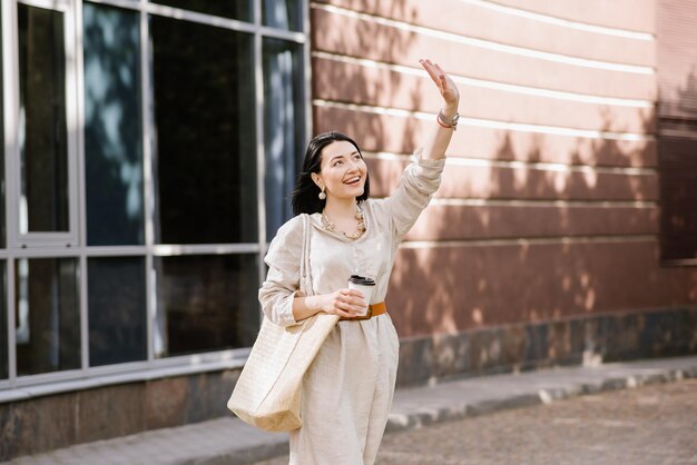 街を歩いてコーヒーを保持しているサングラスとバッグを持つブルネットの若い女性。女性のライフスタイルの肖像画