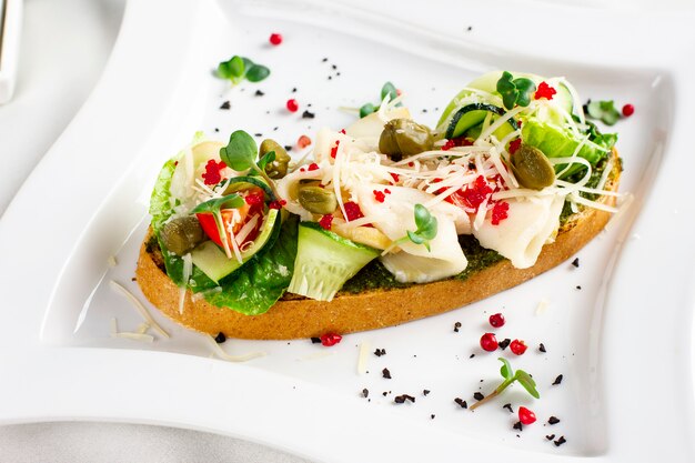 Bruschetta with pesto sauce, fish, fresh vegetables, capers and caviar on white plate