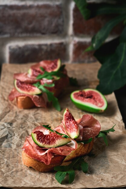 Bruschetta with prosciutto ham, ricotta cheese, arugula and fresh figs on a dark paper