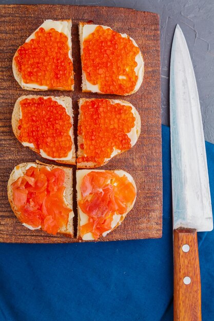 Bruschetta with red caviar and fish on a wooden board next to a knife.