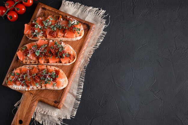 Bruschetta with salmon, dried tomatoes, herbs, cottage cheese, on a black background, wooden board