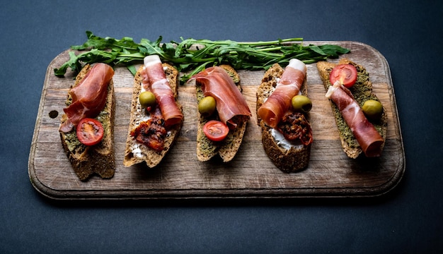 Bruschettas with jamon olives pesto grilled and cherry tomatoes served on wooden board with arugula