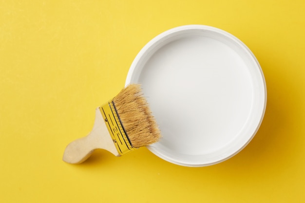 Brush and paint can with white color on a yellow background, top view