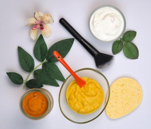 Губка-кисть, ватные диски, желтая натуральная маска для лица в стеклянной миске, листья и цветы куркумы, йогурта