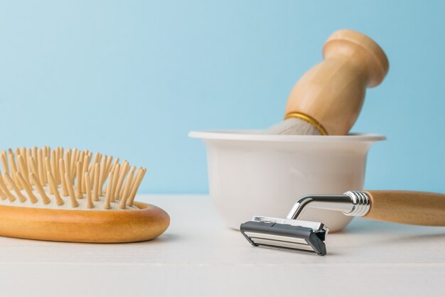 A brush in a white bowl, a razor and a comb on a white table on a blue background.