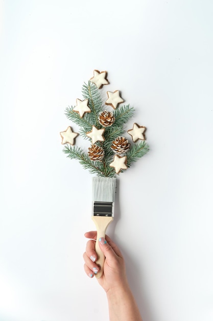 Brush with Christmas tree twigs, natural fir decorated with pine cones and star cookies. Creative simple minimal winter background. Hand with manicured nails holding brush. Winter time DIY renovation.