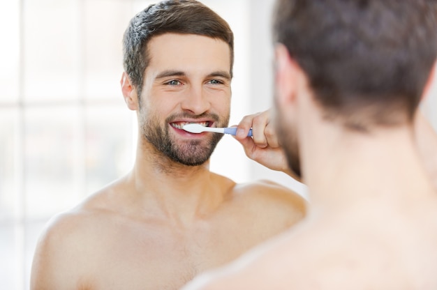 Foto lavarsi i denti al mattino. vista posteriore di un bel giovane uomo con la barba che si lava i denti e sorride mentre sta in piedi contro uno specchio