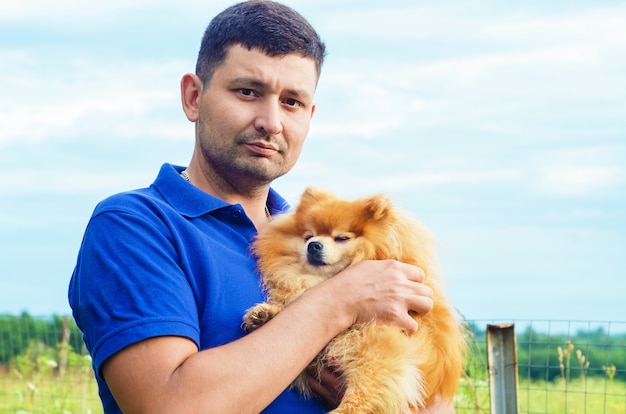 Brutal attractive man smiling and holding pomeranian spitz in hands. owner hugging the dog, spending leisure time together outdoors. pet adoption. friendship of human and animal