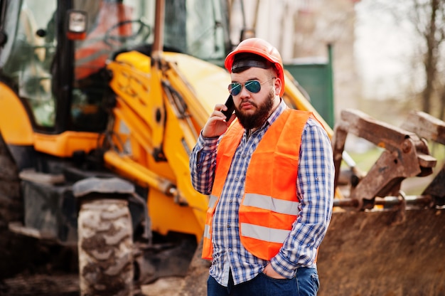 Foto brutal barba operaio uomo vestito operaio edile in casco di sicurezza arancione, occhiali da sole contro traktor con il telefono cellulare a portata di mano.