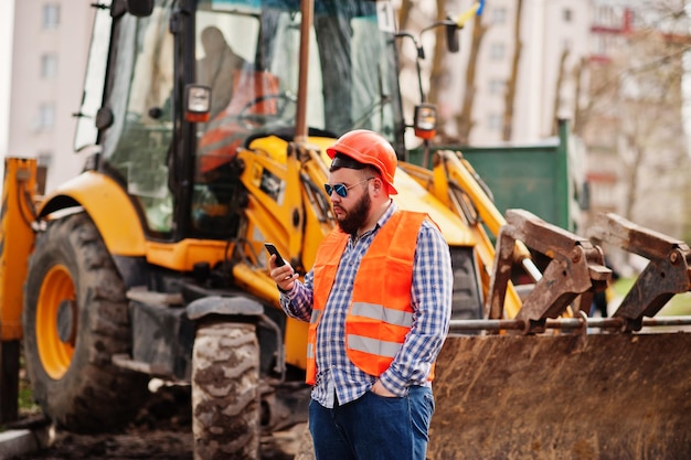 Brutal beard worker man suit construction worker in safety orange helmet sunglasses against traktor with mobile phone at hand