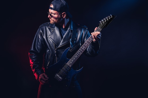Brutal bearded heavy metal musician in leather jacket cap and sunglasses is playing electrical guitar on black 