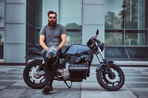 Brutal bearded male in a gray t-shirt and black pants holds a helmet sitting on his custom-made retro motorcycle against a skyscraper, looking at a camera.