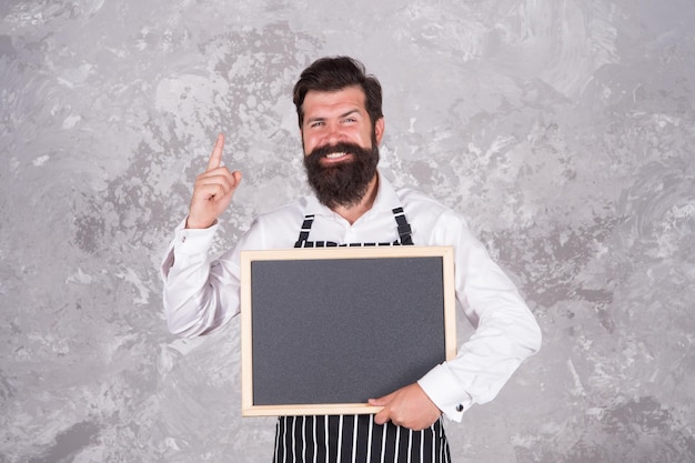 Brutal bearded man chef presenting restaurant menu on empty board for copy space advertising, culinary.