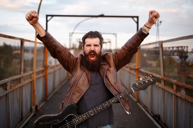 Brutal bearded man a guitarist with a mustache in a brown leather jacket raised his hands and shouts with a guitar