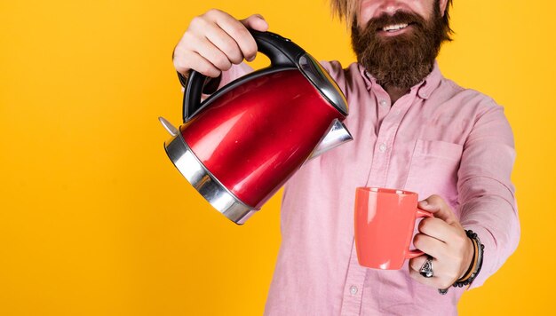 брутальный кавказский хипстер с бородой в рубашке держит электрический чайник, пьет чай.