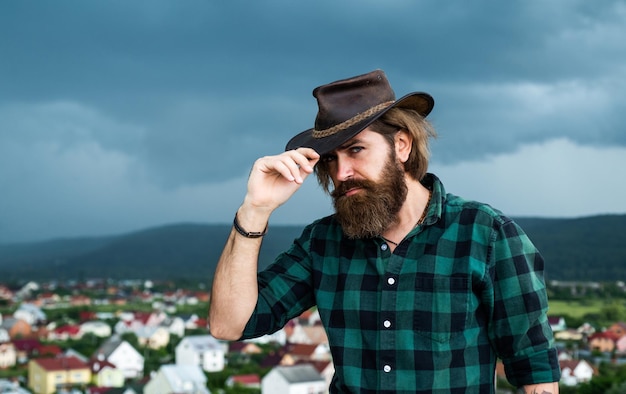 Brutal caucasian hipster with moustache on cloudy sky background. male brutal beauty. confident western guy in checkered shirt. bearded man. mature hipster with beard in cowboy hat. Perfect style.