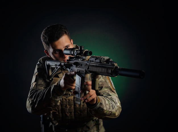 A brutal guy in military airsoft overalls poses with a weapon in his hands on a dark background