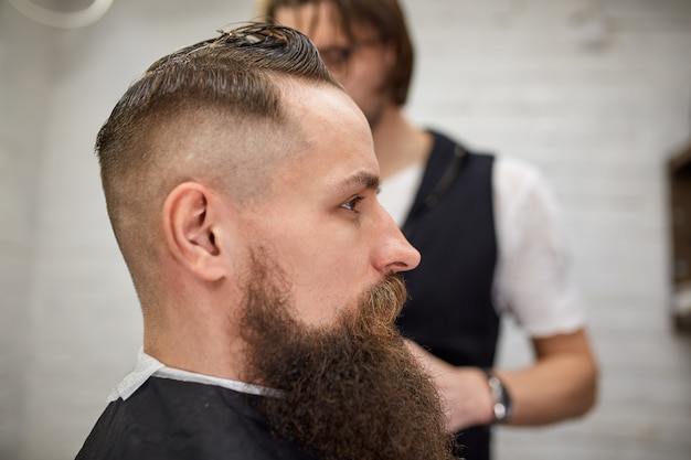 Photo brutal guy in modern barber shop. hairdresser makes hairstyle a man with a long beard. master hairdresser does hairstyle with hair clipper