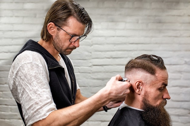 Брутальный парень в современной парикмахерской. Парикмахер делает прическу мужчине с длинной бородой. Мастер-парикмахер делает прическу с помощью машинки для стрижки волос