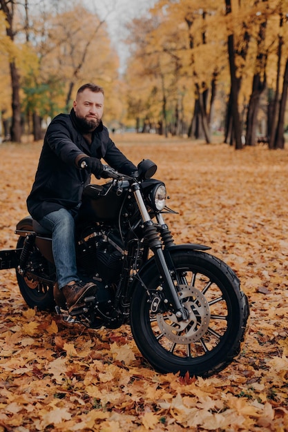 Brutal male biker rides motorcycle during autumn time poses in park with many trees and yellow foliage enjoys driving in open air has tour journey Motorcyclist with own transport outdoor