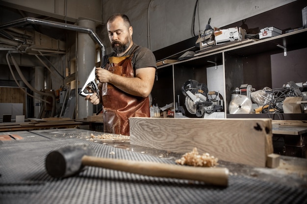 Brutal master carpenter in an apron adjusts plane in workshop