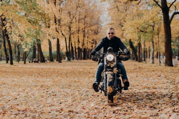 Brutal professional male motorcyclist rides bike wears sunglasses gloves and black jacket has ride through autumnal park beautiful scenery in background with yellow trees and fallen leaves around