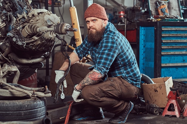 Брутальный татуированный бородатый механик ремонтирует двигатель автомобиля, который поднят на гидравлическом подъемнике в гараже. Станция обслуживания.