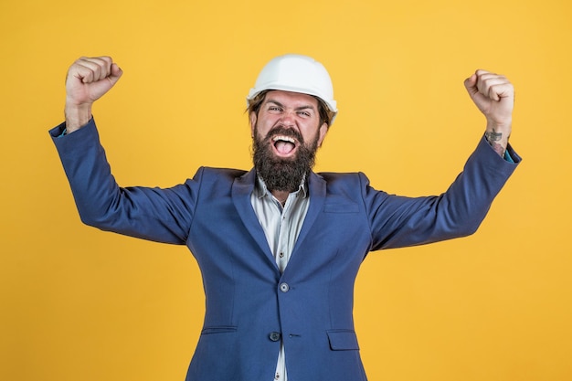 Brutal unshaven guy in formal wear and hard hat on building site, building.