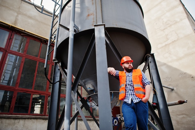 Brutale baard werknemer man pak bouwvakker in veiligheid oranje helm verblijf in de buurt van grote industriële vat.