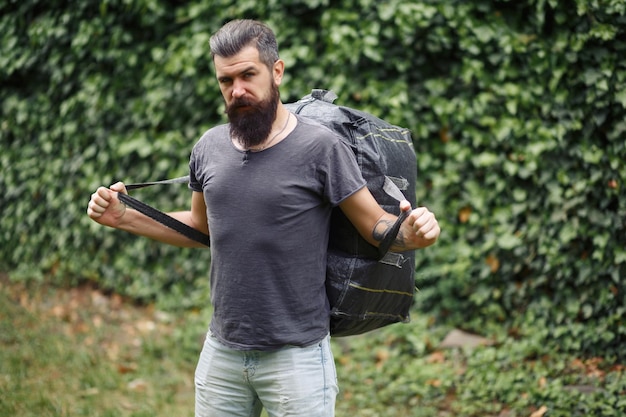 Brutale bebaarde mannelijke koerier in korte broek en een tshirt met een grote grijze tas op zijn schouders in een groen park dakloze niet geschoren man met een grote grijze tas