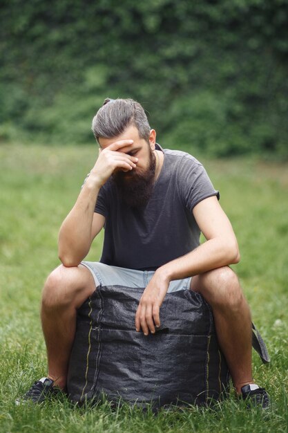 Brutale bebaarde mannelijke koerier in korte broek en een tshirt met een grote grijze tas rust in een groen park dakloze man niet geschoren met dingen in de natuur