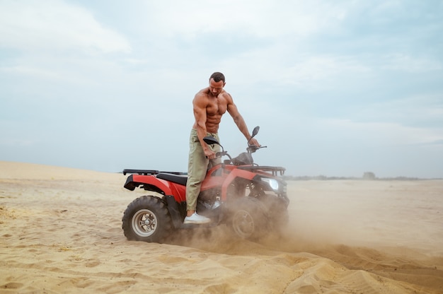 Brutale man met naakt lichaam rijdt op atv in woestijnzand, quadbike-racer. Mannelijke persoon op quad, zandrace op quadbike, duinsafari op warme zonnige dag, 4x4 extreem avontuur, quadrijden